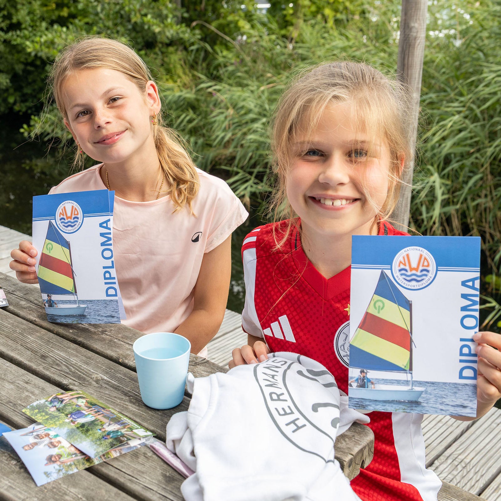 Two young girls holding diplomas outdoors on a wooden table with greenery in the background.