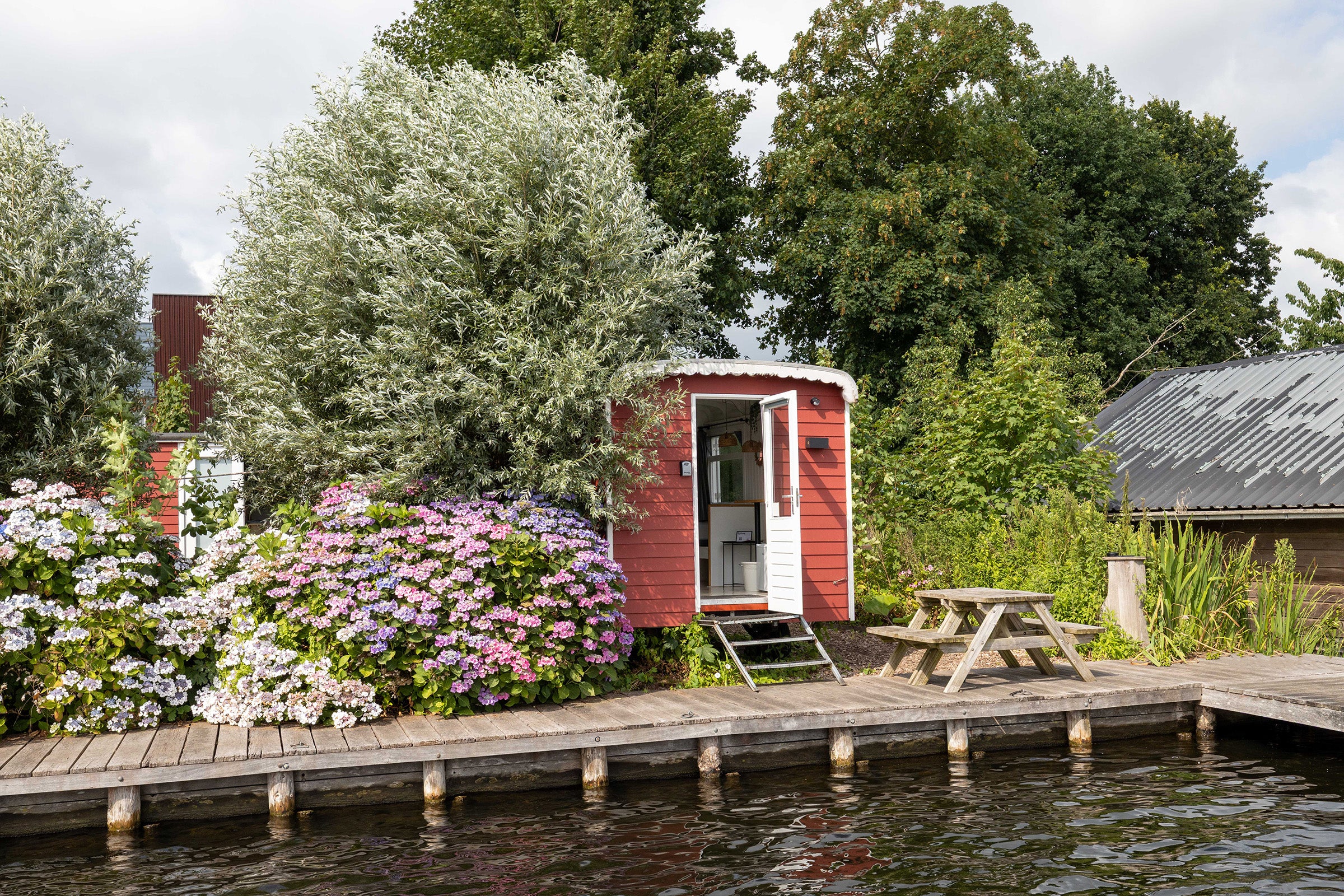 Red shed by a body of water with flowers and a wooden dock.