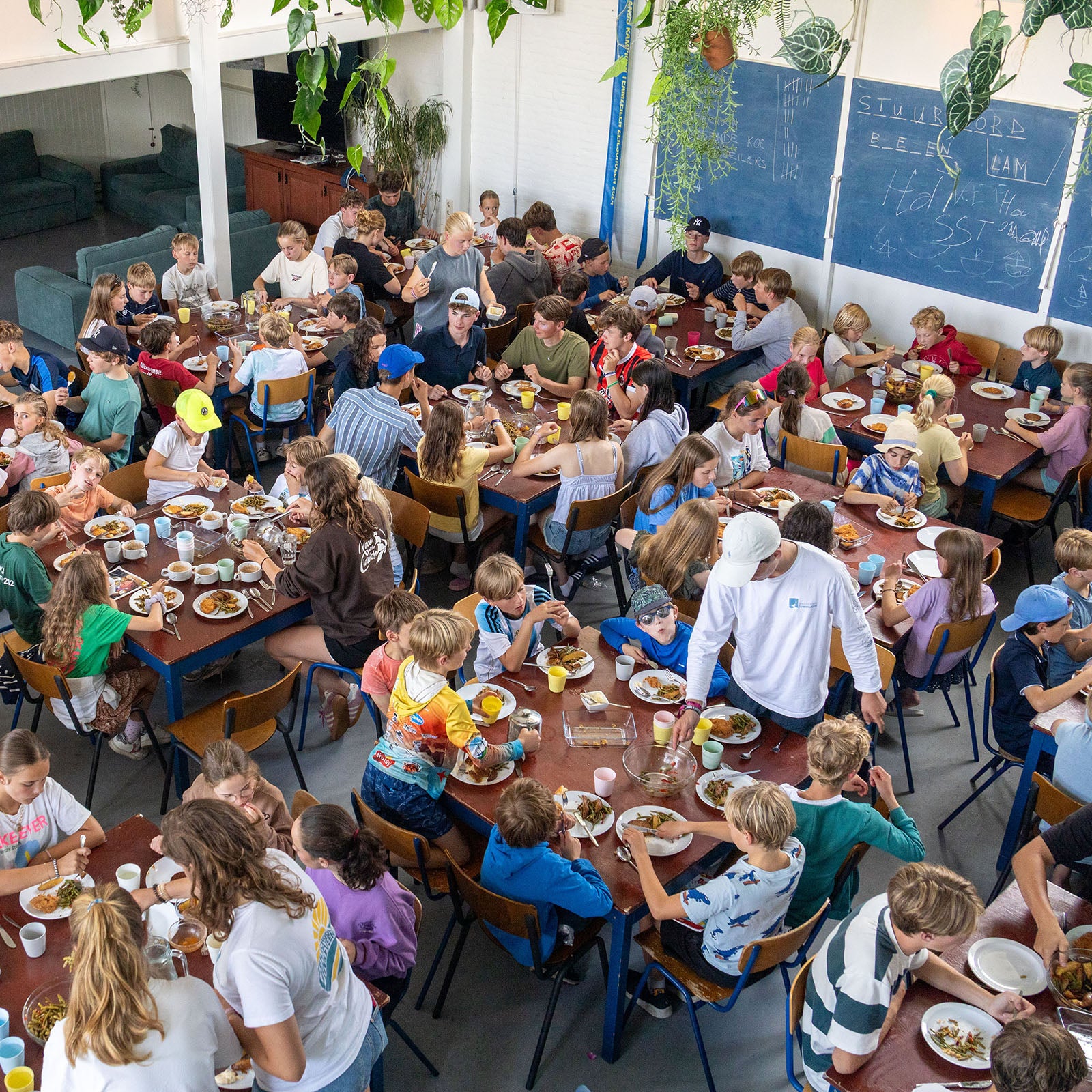 Group of children and adults sitting at tables eating together in a large room with plants and furniture.